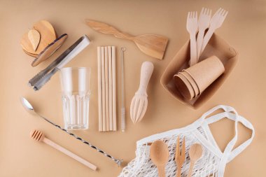 Ahşap çatal bıçak takımı, kağıt tek kullanımlık masa takımı ve bej arka planda bar aletleri. Çevre dostu mutfak aletleri ve ağ torbası. Sıfır atık, plastiksiz konsept. Üst görünüm