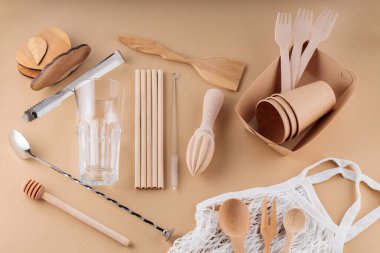 Ahşap çatal bıçak takımı, kağıt tek kullanımlık masa takımı ve bej arka planda bar aletleri. Çevre dostu mutfak aletleri ve ağ torbası. Sıfır atık, plastiksiz konsept. Üst görünüm