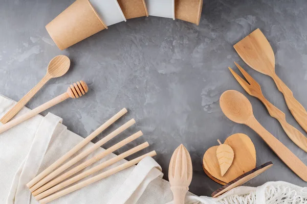 Ahşap masa takımı, kağıt bardaklar ve gri arka planda keten alışveriş torbası. Sıfır atık seti, ağ torbası, bambu kamışları ve ahşap çatal bıçaklar. Çevre dostu kavram