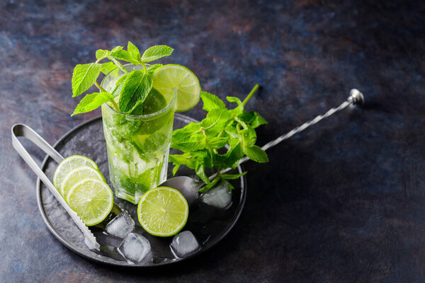 Мохито коктейль, ломтики лайма, мяты и льда на темном фоне. Коктейль мохито, ледяные щипцы и ложка для коктейля на черной тарелке. Вид сверху. Копирование пространства