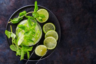 Mojito kokteyli, arkaplanda nane ve limon dilimleri. Siyah bir tabakta yaz kokteyli mojito ve buz maşası. Üst Manzara. Boşluğu kopyala