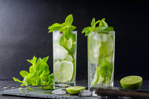 Mojito cocktail, mint, lime slices and ice cubes on a dark background. Cocktail mojito, ice tongs and cocktail spoon on slate board. Copy space