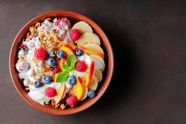 Yoğurtlu granola kahvaltısı, yabanmersini, ahududu, muz ve şeftali. Yoğurtlu sağlıklı kahvaltı, pişmiş tahıl ve kil tabakta meyve. Üst Manzara. Boşluğu kopyala