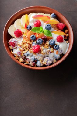 Yoğurtlu granola kahvaltısı, yabanmersini, ahududu, muz ve şeftali. Yoğurtlu sağlıklı kahvaltı, pişmiş tahıl ve kil tabakta meyve. Üst Manzara. Boşluğu kopyala