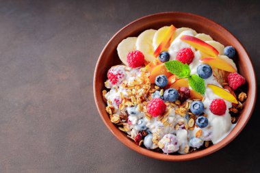 Yoğurtlu granola kahvaltısı, yabanmersini, ahududu, muz ve şeftali. Yoğurtlu sağlıklı kahvaltı, pişmiş tahıl ve kil tabakta meyve. Üst Manzara. Boşluğu kopyala