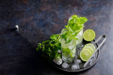 Two glasses of mojito cocktail, lime slices, mint and ice on dark background. Refreshing mint cocktail mojito, ice tongs and cocktail spoon on black plate