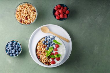 Greek yogurt with raspberries, blueberries and granola on a green background. Bowl with healthy breakfast on white plate with wooden spoon. Top view. Copy space
