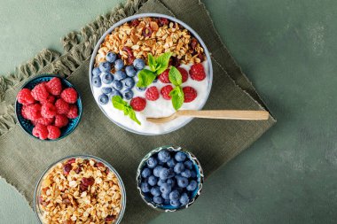 Homemade yogurt bowl with raspberry, blueberry and granola on a green surface. Bowl with yogurt, granola and berries on a green linen napkin. Healthy food concept