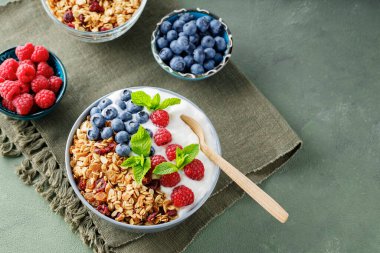Homemade yogurt bowl with raspberry, blueberry and granola on a green surface. Bowl with yogurt, granola and berries on a green linen napkin. Healthy food concept