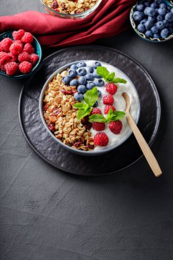 Breakfast cereal granola with berries and greek yogurt on a dark background. Bowl with yogurt, granola and fresh berries on a black plate. Healthy breakfast with wooden spoon