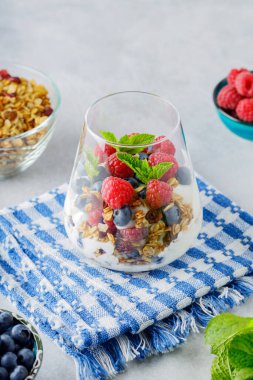 Glass with granola, yogurt, fresh raspberries, blueberries and mint. High protein and diet breakfast on a white-blue napkin. Healthy food concept