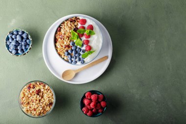 Greek yogurt with raspberries, blueberries and granola on a green background. Bowl with healthy breakfast on white plate with wooden spoon. Top view. Copy space