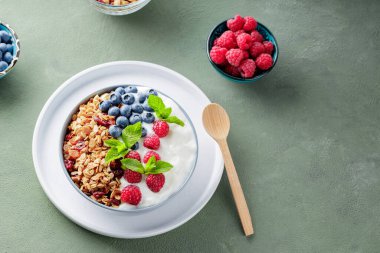Greek yogurt with raspberries, blueberries and granola on a green background. Bowl with healthy breakfast on white plate with wooden spoon. Top view. Copy space