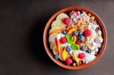 Yoğurtlu granola kahvaltısı, yabanmersini, ahududu, muz ve şeftali. Yoğurtlu sağlıklı kahvaltı, pişmiş tahıl ve kil tabakta meyve. Üst Manzara. Boşluğu kopyala