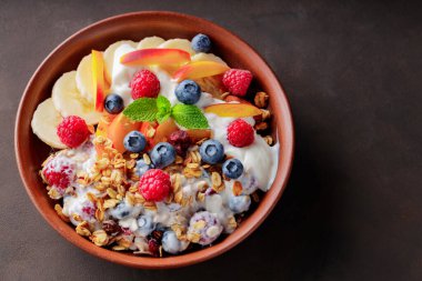 Yoğurtlu granola kahvaltısı, yabanmersini, ahududu, muz ve şeftali. Yoğurtlu sağlıklı kahvaltı, pişmiş tahıl ve kil tabakta meyve. Üst Manzara. Boşluğu kopyala
