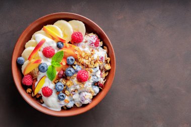 Yoğurtlu granola kahvaltısı, yabanmersini, ahududu, muz ve şeftali. Yoğurtlu sağlıklı kahvaltı, pişmiş tahıl ve kil tabakta meyve. Üst Manzara. Boşluğu kopyala