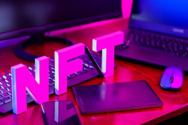 3 boyutlu bir yazıcıya basılmış 3 boyutlu harflerden yapılmış NFT yazıtları. İş yerinde grafiksel tablet, akıllı telefon ve bilgisayar donanımlı NFT harfleri var. Pembe-mavi ışıkta iadesi mümkün olmayan jeton.