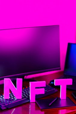 Pembe neon ışıklı NFT yazıtları. İş yerinde grafiksel tablet, akıllı telefon ve bilgisayar donanımlı NFT harfleri var. Boşluğu kopyala