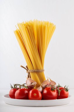 İple bağlanmış bir demet çiğ spagetti. Vişneli domatesli kuru makarna ve beyaz tabakta sarımsak. Yakın plan.