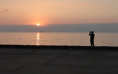 Erkek silueti telefonda, gün doğumunda, deniz kıyısında fotoğraf çekiyor..