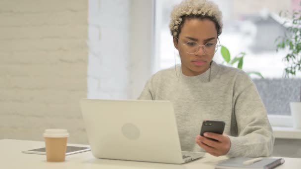 Mujer africana enojada hablando en Smartphone mientras usa el ordenador portátil en la oficina — Vídeos de Stock