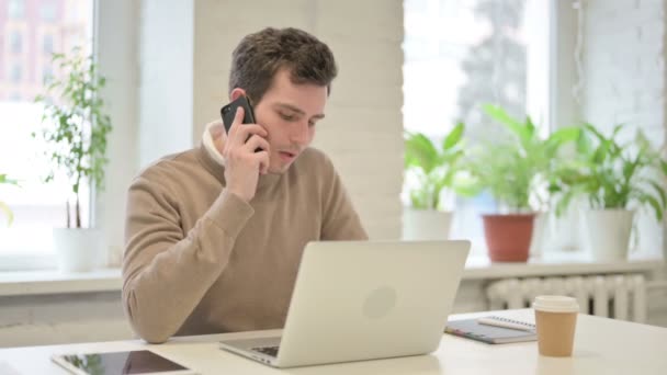 Чоловік розмовляє на смартфоні під час використання ноутбука в офісі — стокове відео