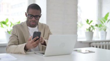 Ofiste dizüstü bilgisayar kullanırken akıllı telefon kullanan Afrikalı adam 