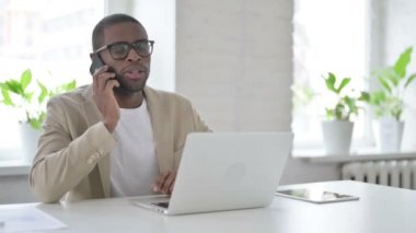 Ofiste dizüstü bilgisayar kullanırken akıllı telefondan konuşan Afrikalı adam.