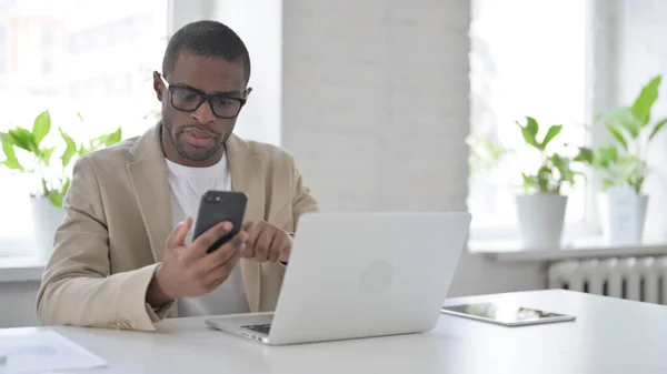 African Man using Smartphone while using Laptop in Office — Stockfoto
