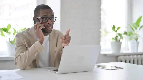 Arg afrikansk man talar på smartphone när du använder laptop i Office — Stockfoto
