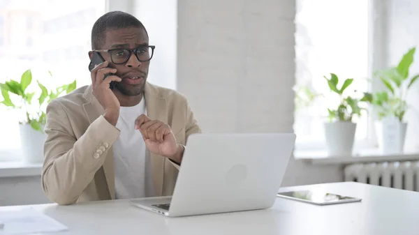 African Man Talking on Smartphone while using Laptop in Office — Stockfoto