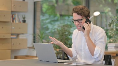 Modern Ofiste Dizüstü bilgisayar kullanırken Akıllı Telefonla Konuşan Kızgın Adam