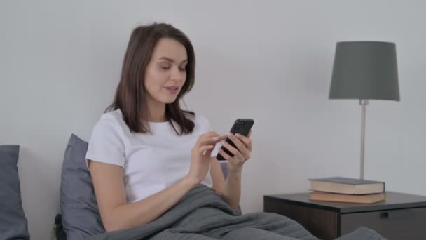Женщина празднует успех на смартфоне в постели — стоковое видео