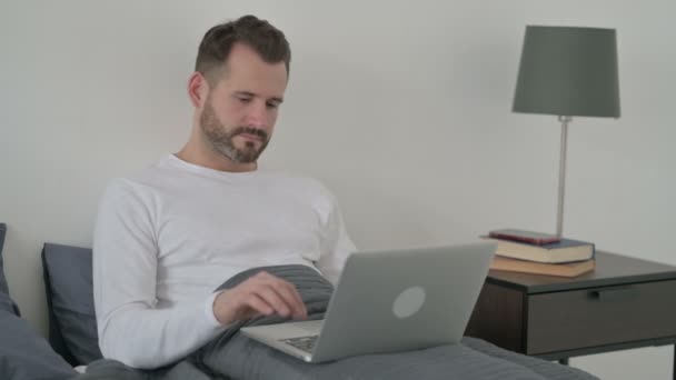 Man met laptop glimlachend naar de camera in bed — Stockvideo