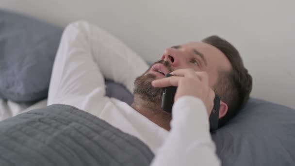 Мужчина разговаривает по телефону, лежа в кровати — стоковое видео