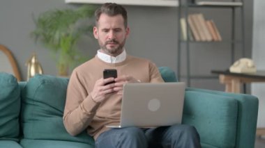 Kanepede akıllı telefon kullanan dizüstü bilgisayarlı bir adam. 