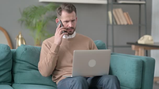 Dühös férfi laptoppal beszél okostelefonon a kanapén — Stock videók