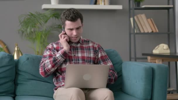 Homem irritado com laptop conversando no smartphone no sofá — Vídeo de Stock