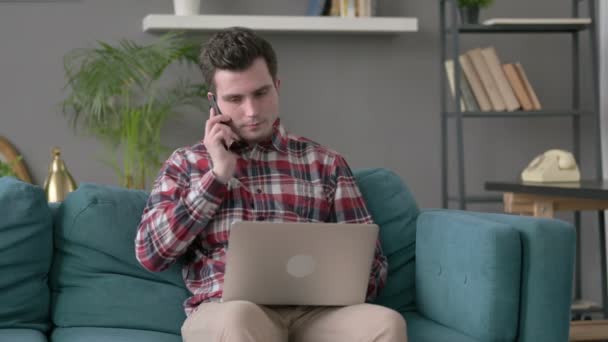 Человек с ноутбуком разговаривает на смартфоне на диване — стоковое видео