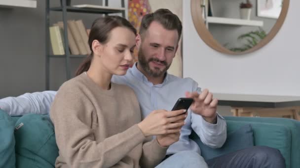 Portret van een paar vieren succes op de smartphone, Zittend op de bank — Stockvideo