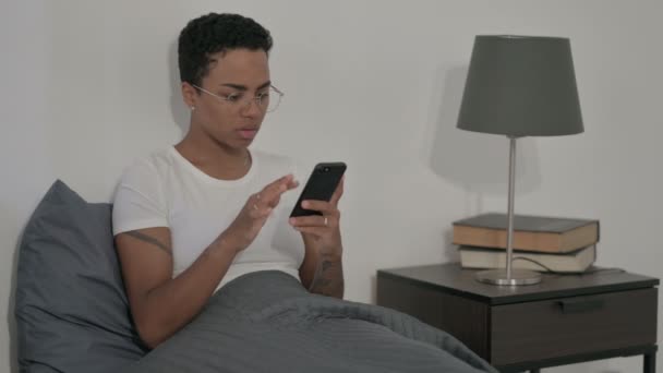Mujer africana reaccionando a la pérdida en el teléfono inteligente en la cama — Vídeos de Stock