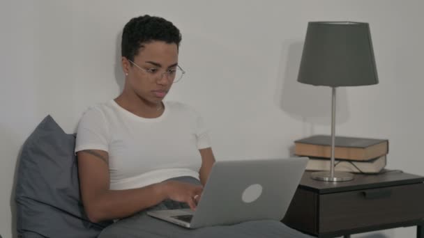 Mujer africana sonriendo a la cámara mientras usa el ordenador portátil en la cama — Vídeos de Stock