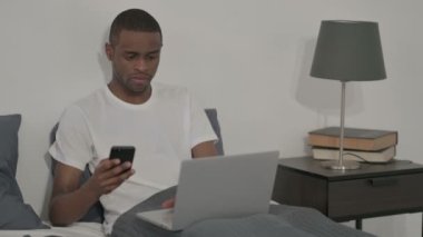Akıllı telefon kullanan, yatakta oturan, dizüstü bilgisayarı olan Afrikalı adam.