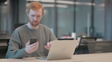 Ofiste dizüstü bilgisayar kullanırken akıllı telefon kullanan genç adam. 