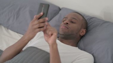 Afrikalı adam yatakta uyurken akıllı telefon kullanıyor. Kapat.