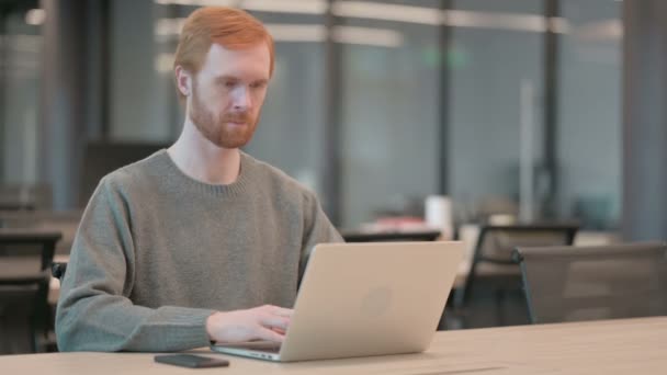 Молодой человек улыбается в камеру во время использования ноутбука в офисе — стоковое видео