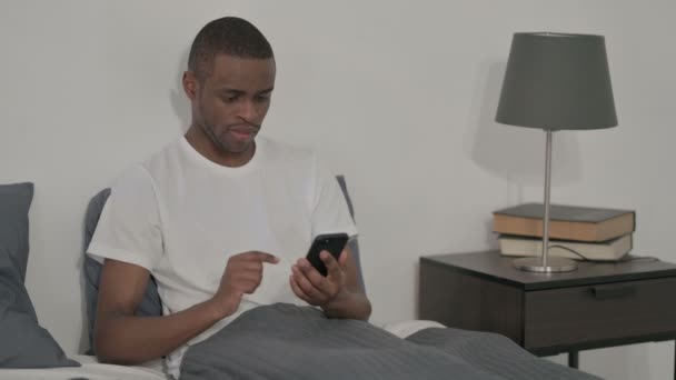 Afrikaner feiert Erfolg auf Smartphone, im Bett sitzend — Stockvideo