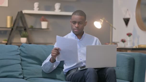 Hombre africano con computadora portátil reaccionando a la pérdida de documentos, sofá — Vídeos de Stock