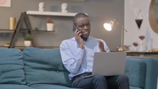 Hombre africano enojado hablando en Smartphone mientras usa el ordenador portátil en el sofá — Vídeos de Stock