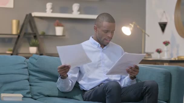 Hombre africano celebrando el éxito mientras lee documentos en el sofá — Vídeos de Stock